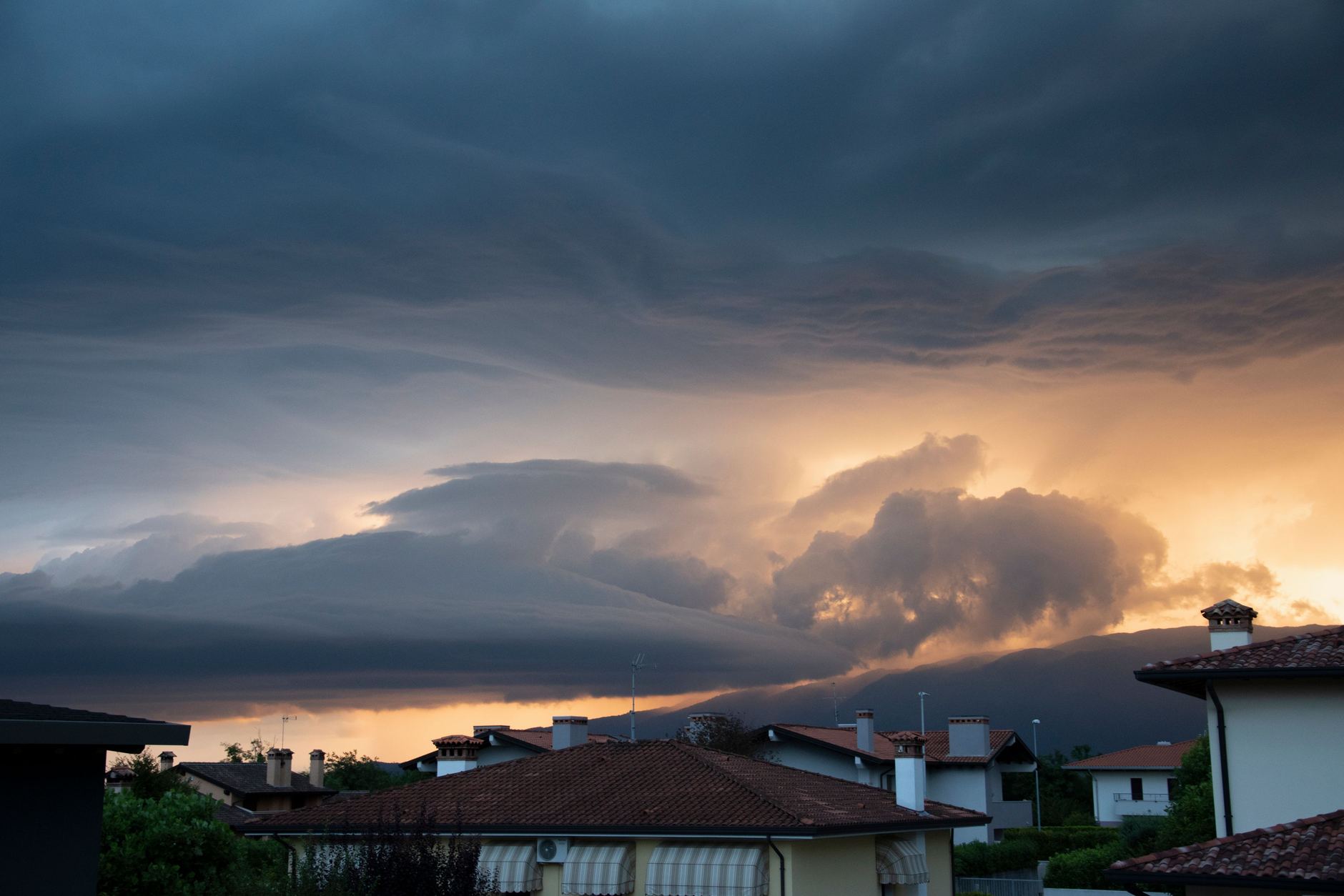 A dark shelf cloud from a severe thunderstorm rolling in over a row of suburban rooftops at dusk