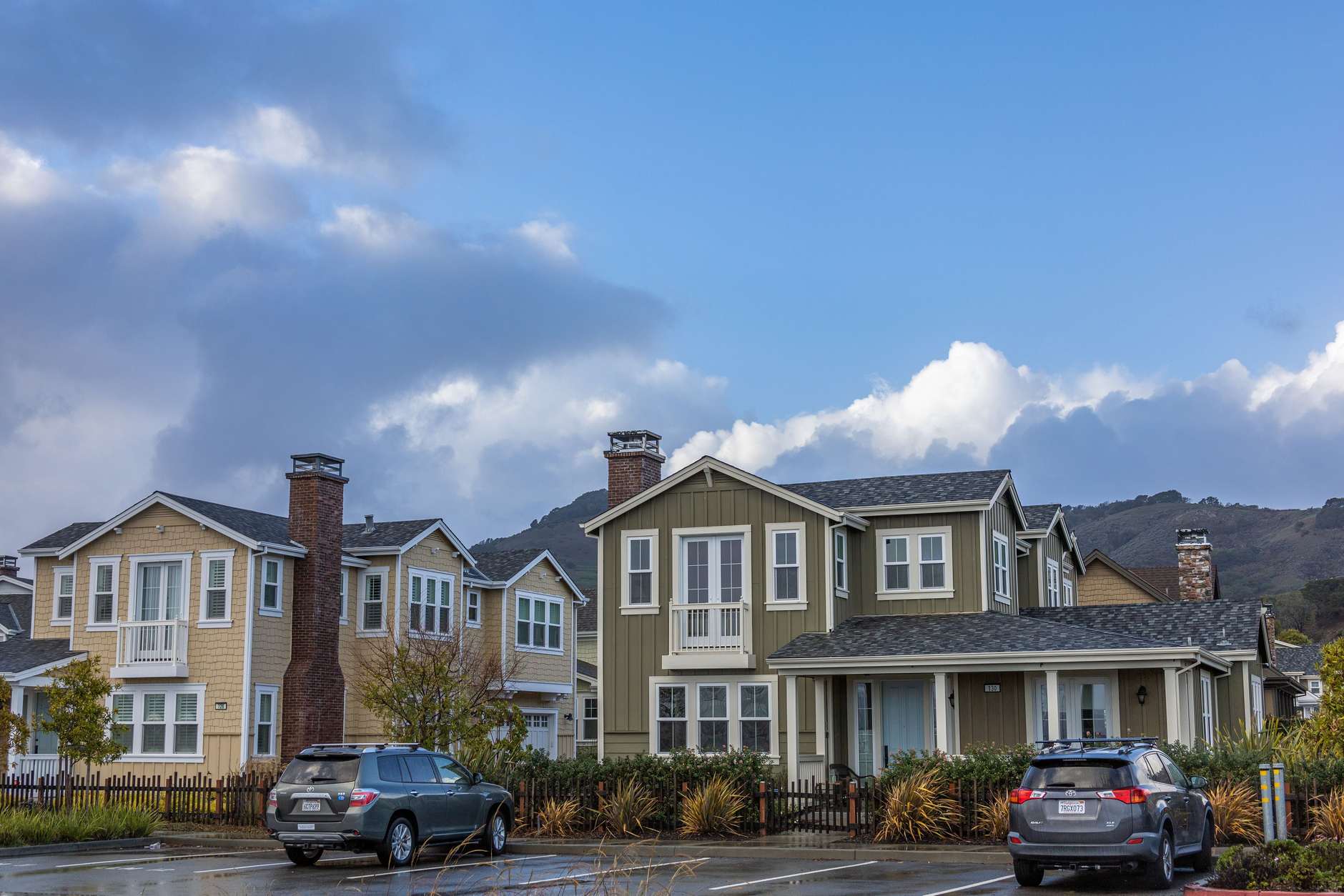 A quiet residential street lined with two-storey houses and parked cars under a blue sky