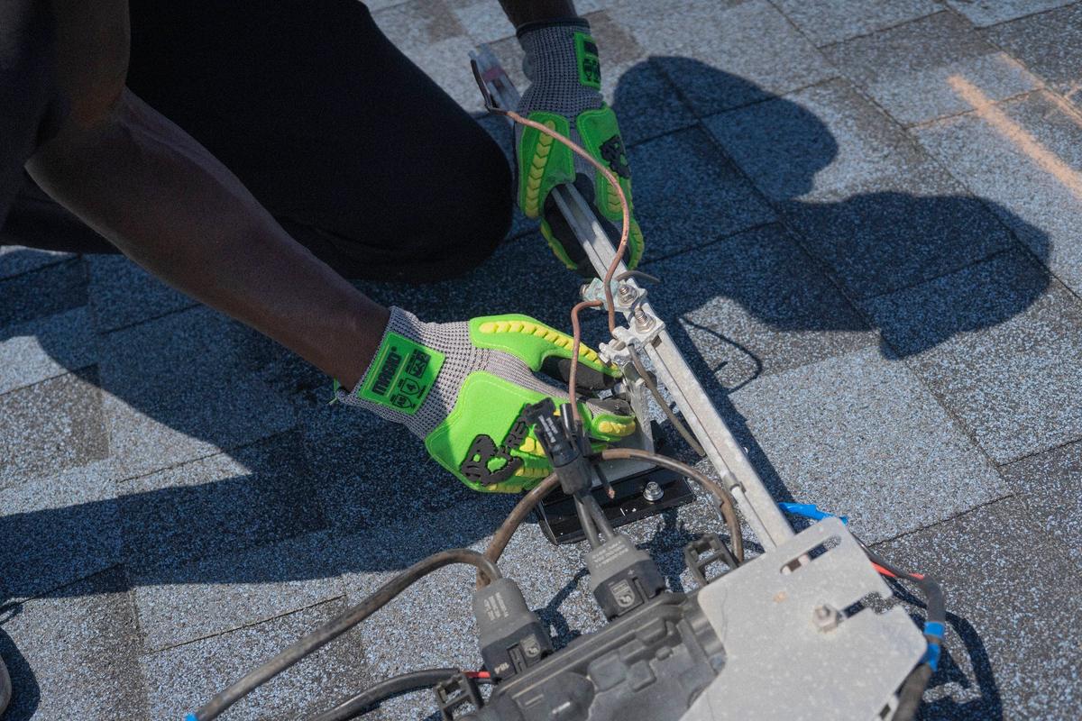 A roofer in work gloves running a nail gun along a fresh row of asphalt shingles
