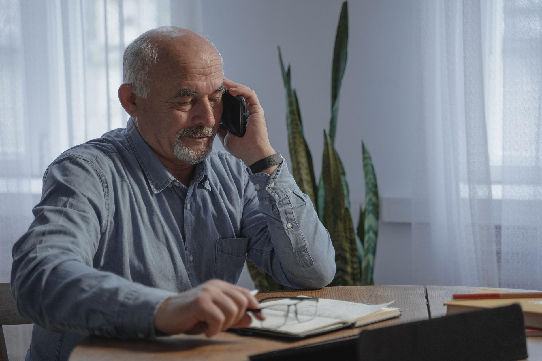 An older business owner on a phone call at his desk, writing notes in a notebook