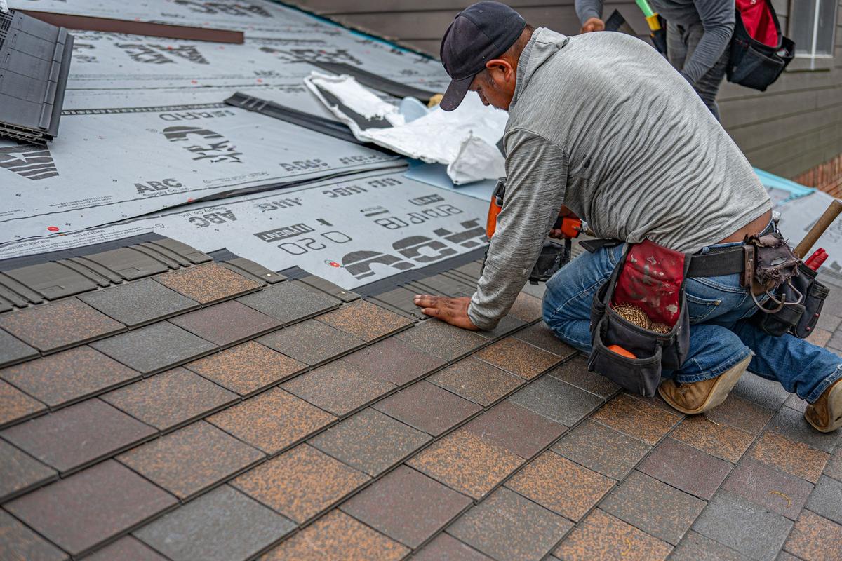 A roofer kneeling on a pitched roof laying new architectural shingles over underlayment